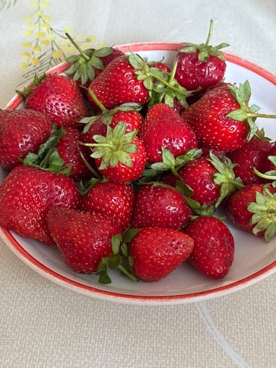 A close-up, overhead shot shows a white ceramic bowl filled with ripe, red strawberries. The bowl has a thin red trim around its rim. The strawberries are of various sizes and are clustered together, with some of their green leafy tops visible. The bowl is placed on a textured, light-colored tablecloth that has a subtle floral pattern. The background is slightly out of focus, but the tablecloth is clearly visible. There are no people or animals in the image. The lighting suggests it is daytime, and the scene is indoors, likely in a home setting. The image does not provide any information or context related to the city of Новый Кинер, Russia.