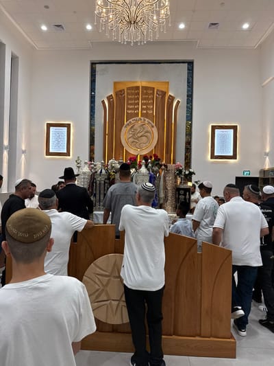 The image depicts the interior of a contemporary synagogue in Beer Yaaqov, Israel, during a gathering of approximately 20-30 males. The focal point is a large, ornate wooden Ark (Aron Kodesh) centered against the rear wall. The Ark features a central circular medallion with a swirling, abstract carved design and the Hebrew inscription "שיויתי ה' לנגדי תמיד." Above the medallion, two vertical panels display Hebrew text, likely from the Ten Commandments. The Ark is flanked by textured dark blue/grey panels.

Directly in front of the Ark, on a raised platform, are numerous Torah scrolls, each enveloped in elaborate silver-colored or embroidered mantles and topped with decorative finials (Rimonim). Several floral arrangements, primarily red and white, are interspersed among the scrolls.

The synagogue interior features white walls and a high ceiling with recessed lighting and a large, multi-tiered crystal chandelier. Two illuminated, framed plaques displaying dense Hebrew text are mounted on the side walls.

The gathered individuals are predominantly adult males, with some younger individuals also present. All visible men and boys are wearing kippahs (head coverings) of various designs and colors. Their attire is mostly casual, including t-shirts and collared shirts, though one man in the mid-ground wears a black suit and fedora. All individuals are standing, generally oriented towards the Ark and Torah scrolls. In the immediate foreground, a light-colored wooden barrier or partition with carved circular and linear decorative elements is visible. An "EXIT" sign, in both Hebrew and English, is visible on the far right wall.