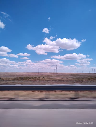 The image captures a vast, flat landscape under a bright blue sky with scattered white clouds, characteristic of the steppes near Dams, Kazakhstan. The foreground is dominated by a blurred representation of a road or ground, suggesting movement. A metallic guardrail runs horizontally across the mid-ground, separating the road from the arid terrain. Beyond the guardrail, the landscape unfolds as a dry, brown plain dotted with sparse vegetation and a few scattered power poles. In the distance, several tall power pylons are visible, indicating infrastructure within this remote area. The sky is a vibrant blue, with puffy white clouds creating a dynamic overhead view. The timestamp in the bottom right corner reads "20 июля 2026 г., 11:57", indicating the date and time the photograph was taken.