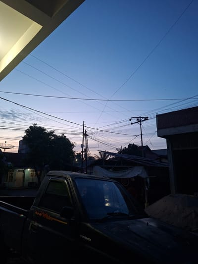 The image depicts a street scene at dusk in the village of Meunasah Dayah, Bireuen, Indonesia. The sky is a gradient of deep blue to pale purple, with numerous power lines crisscrossing the frame. In the foreground, the hood and driver's side of a dark-colored pickup truck are visible. The truck has "Pick-Up" and "PANTHER" text on its body. To the left, a portion of a building's overhang is illuminated. In the middle ground, a large tree with dark foliage stands near a house with lights on inside and a satellite dish on its roof. A utility pole with many wires is also prominent. To the right, a brick building under construction and a pile of sand are visible. The overall scene is one of a quiet, developing residential area as day transitions into night.