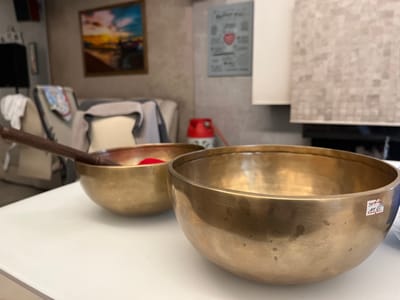 The image shows two brass singing bowls on a white table. The bowl on the left has a wooden mallet inside. The bowl on the right has a small white price tag on its side. The background includes a painting, a wall decoration, a sofa, a small red gas cylinder, and a white cabinet with a tiled backsplash. The specific location in the city of Patrony, Russia cannot be determined from the image.