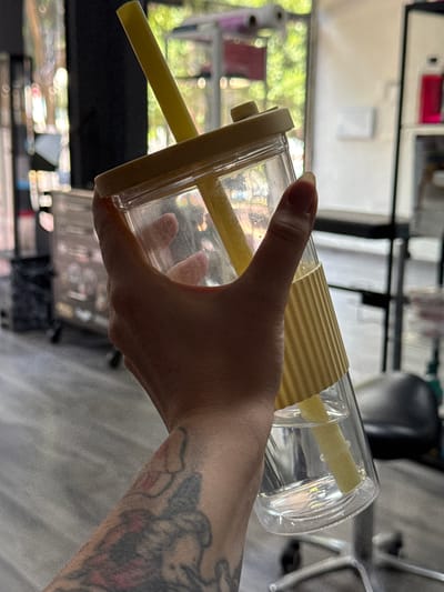 A hand with a tattoo on its arm holds a clear plastic cup with a yellow straw. The cup is angled, and the straw extends from the cup and out of frame to the left. A yellow corrugated band wraps around the middle of the cup. The background is blurred, showing greenery and what appear to be shelves with various items. The lighting suggests it is daytime. The setting appears to be indoors, possibly a home or a shop, with a window or large opening allowing light and a view of the outside. The tattoo on the arm is detailed, featuring shading and colors. The person's fingernail on the thumb is visible and appears to be painted.