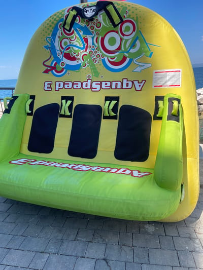 An inflated yellow and green tube-shaped watercraft rests on a paved walkway with the sea and a rocky shoreline visible in the background under a clear blue sky. The watercraft, which appears to be a towable raft for multiple people, has the word "AquaSpeed 3" printed in white with a red outline on its side. There are also black cushioned seats and yellow straps with the letter "K" on them. A warning label is affixed to the upper right portion of the watercraft. The location is Crveni Vrh, Croatia.