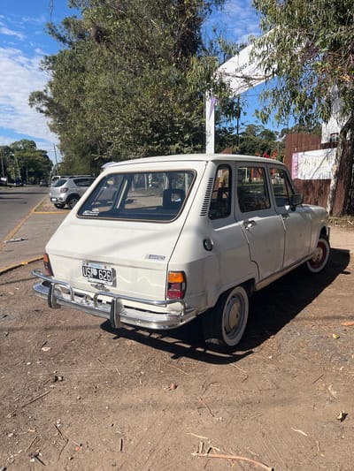 A white vintage Renault 6 GTL automobile, bearing Argentine license plate USP 626, is parked on an unpaved, light brown dirt surface. The vehicle features chrome bumpers, factory hubcaps, and white wall tires; the rear right wheel rim is distinctly red. A partial view of an individual's torso and arm, clothed in an olive-green shirt, is visible through the open front passenger window.
The vehicle is positioned at the entrance to the Museo del Transporte Carlos Hillner y Nicolorich in Quilmes Oeste, Argentina. The entrance is demarcated by a white archway with a partial inscription of "Museo del Transporte," situated amidst dense green trees and a brown wooden fence. To the immediate left of the parked car, an asphalt street with a yellow-curbed sidewalk is visible, featuring several parked vehicles, including a silver crossover model further down. Utility poles are present along the street. The scene is illuminated by natural daylight under a clear blue sky scattered with white clouds. The immediate ground is characterized by uneven dirt, intermingled with small stones, twigs, and dry leaves, and exhibits faint tire tracks.
