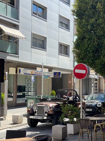 Here is a detailed description of the image:

**General Description:**
The image shows a street scene, apparently in the city of Llíria, Spain. The photo is oriented vertically, showing urban elements, vehicles, signage, and a person.

**People:**
- There is a person visible behind the steering wheel of an open-top vehicle (likely a Jeep).

**Subjects & Objects:**
- **Vehicles:** An open-top Jeep-like vehicle is prominent. Part of another car (a Mini Cooper with the license plate "645 MKS") is also visible.
- **Signage:** A prominent “no entry” sign is visible, as well as signs for “Llíria Invests,” a real estate and urban services company. Other signs point to tourist routes and Ermita de Santa Barbara.
- **Building:** A modern building with multiple windows is visible in the background. There is an awning over the building's lower floor.
- **Street furniture:** Chairs and a table from an outdoor café are visible, suggesting the street has restaurants or cafes.

**Location:**
Based on the “Llíria Invests” signage and the information on signs, it is likely in the city of Llíria, Spain. The Ermita de Santa Barbara sign further corroborates this location.

**Actions & Interactions:**
- The Jeep is parked, possibly blocking part of the sidewalk near the outdoor café seating.

**Environment:**
- It is a sunny day. The photo is taken on a street that includes residential or commercial buildings and parking. A tree overhangs the scene, adding a touch of greenery.