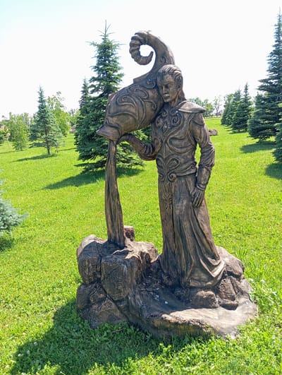 The image shows a statue of a man pouring liquid from a large, ornate jug. The statue is made of a dark, rough material that resembles wood or stone and is placed outdoors on a grassy lawn. The man is wearing a flowing robe and intricate armor, with a serious expression on his face. He is holding the jug with his right hand, and the liquid is pouring down onto a rocky base. The jug itself has a coiled, horn-like handle. The background consists of lush green grass, several evergreen trees, and a clear blue sky. The lighting suggests it is a sunny day. The statue is located in the city of Il'ich, Russia.