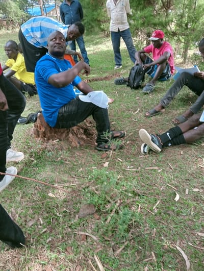 The image captures an outdoor gathering of at least eight adult males in a grassy, partially shaded area within Lwandeti ward, Kenya. The ground is covered with green grass, patches of bare soil, and scattered dry leaves and small stones.
In the foreground-center, a male subject with a dark complexion and stout build is seated on a rough tree stump. He wears a blue short-sleeved t-shirt, black pants, and has a white baseball cap resting on his lap. His head is tilted back, and he is looking upwards and to his right. Directly behind him, another male subject, wearing a light blue and white striped t-shirt and dark pants, is leaning forward with his gaze directed towards the seated man.
To the seated man's left, a partially visible male in a yellow t-shirt is present. In the mid-ground to the right, a male subject wearing a red baseball cap, a pink t-shirt, and patterned shorts is seated on the ground, interacting with a black backpack that has a microphone attached to its strap. To his immediate right, another male subject is seated on the ground, holding what appears to be a dark tablet or notebook.
In the background, two additional male subjects are standing; one in a dark blue shirt and pants, and another in a light-colored shirt and blue jeans. Partial views of additional legs and footwear, including white sneakers and dark sandals, are visible in the immediate foreground. The environment includes mature trees in the background, with a glimpse of a structure featuring a blue roof further in the distance. The scene indicates a casual outdoor meeting or discussion among the individuals.