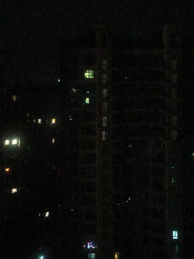 The image shows a dark nighttime scene featuring the exteriors of several buildings, illuminated by lit windows. The buildings appear to be residential or office structures, with multiple stories and visible windows, some of which emit light in various colors, including yellow, white, and green. The overall environment is dark, suggesting an urban setting at night. There are no visible people or specific activities depicted, only the static architecture and its illuminated windows. The image appears to have been taken from a distance, possibly from another building or a vantage point overlooking the cityscape.