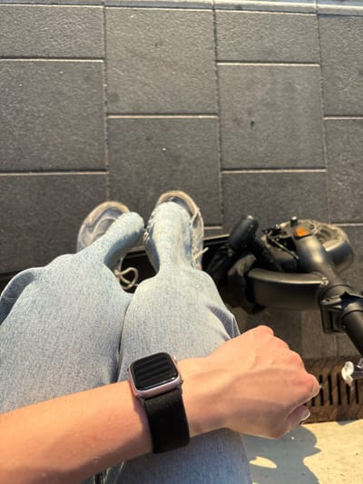 The image shows a first-person perspective of someone sitting on a scooter, with their legs and wrist visible. The person is wearing light blue jeans and white sneakers. On their left wrist, a smartwatch with a dark band is visible. The scooter, which appears to be an electric scooter, is dark gray or black and is positioned in front of the person.
The ground is paved with rectangular gray tiles, suggesting an outdoor urban or suburban setting. A metal grate is visible to the right of the scooter, casting a shadow on the pavement, indicating it is daytime. The lighting suggests it is either morning or late afternoon, with the sun casting long shadows. The overall mood is casual and relaxed, with the subject appearing to be taking a break or preparing to ride. No text is visible in the image. The location context is Elsenfeld, Germany.