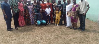A large group of people stands on a grassy lawn. Many adults wear colorful, patterned clothing. There are also several children present, some standing and others being held or posed with by adults. One man is kneeling and holding up a phone, presumably taking a group photo. The background features a light-colored building or structure.
