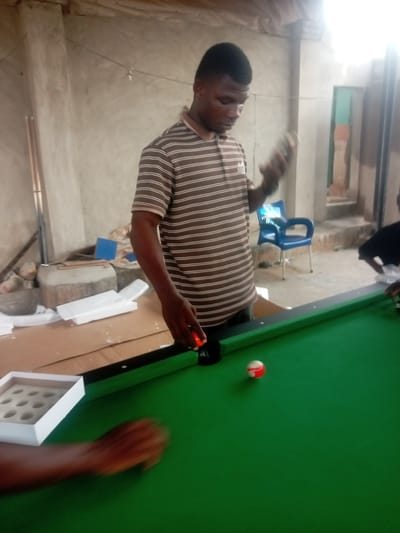 The image depicts a man standing at a green billiard table in what appears to be an indoor/outdoor space in Umuokpu, Nigeria. The man is wearing a striped brown and white shirt and is holding a billiard ball in his right hand. He is focusing on positioning a red ball on the table. Next to the table, there's a white tray with compartments, likely for holding the billiard balls. Another individual's arm, possibly holding a billiard cue, is visible on the left edge of the frame. The background suggests a simple, possibly unfinished construction, with exposed walls and a view to an outdoor area with a blue plastic chair. There's also debris and packing materials in the background, suggesting the billiard table may have been recently set up.