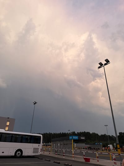 This media file depicts an outdoor scene, likely a bus stop or parking area, in Chociule, Poland. The sky is filled with dramatic clouds, suggesting either dawn or dusk, with warm, light tones in the upper left portion transitioning to darker, more overcast conditions towards the right. Two tall light poles with multiple fixtures are visible, indicating the area is illuminated, possibly for evening use.
In the foreground, a white bus is parked, its side facing the viewer. The bus has tinted windows and a visible rear wheel. To the right of the bus, a sturdy barrier made of white and red poles separates the parking area from what appears to be a pathway or roadway. Beyond the barrier, a line of people, some wearing orange vests, are visible, suggesting an organized event or transportation hub. Further in the background, a dense line of trees borders the scene.
Several signs are present. One prominent blue sign with white text and a bus icon reads "Deszczno," likely indicating a destination or route. Another sign with a "6" and some other illegible text is visible on a yellow pole.
The overall impression is a functional public space at the edge of a wooded area, likely during a transitionary time of day with overcast weather. The presence of people in vests and the bus suggests a moment of transit or waiting.