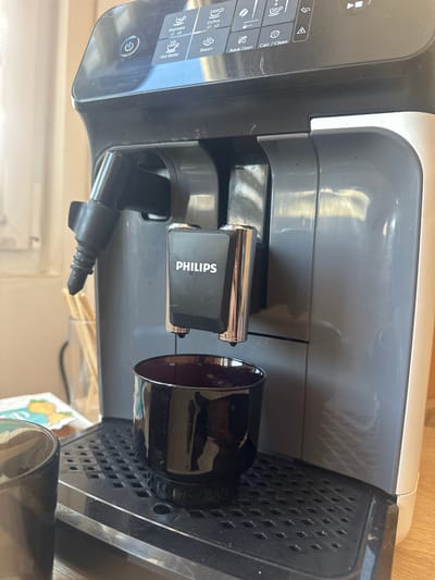 The image shows a Philips automatic espresso machine in a domestic setting. The machine is grey and black, with a digital display on top showing various coffee options like "Espresso", "Coffee", "Hot Water", "Steam", "Aqua Clean", and "Calc/Clean". A black espresso cup is positioned under the dispensing spout, and another partially visible black cup is to the left. A glass of what appears to be wooden stirrers is behind the machine. The setting is likely a kitchen or dining area, with a blurred background that suggests an indoor environment. The image does not provide any information about the location being Orlivka, Ukraine.