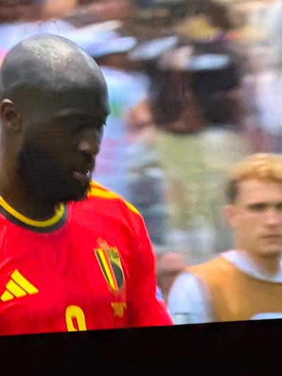 The image shows a close-up of a man, likely a soccer player, wearing a red jersey with yellow trim and a crest on the left side of his chest. The crest features black, yellow, and red elements. A yellow Adidas logo is visible on the right side of his chest. He has dark skin, a shaved head, and a beard. He is looking down and to his right. In the background, another person with blond hair and wearing a yellow and white vest is visible, out of focus. The scene appears to be a sporting event, possibly a soccer match. The location in the city of Bykovo, Russia is not identifiable from the image.
