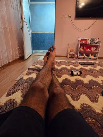 The image shows a first-person perspective of a person's legs and feet resting on a patterned rug in what appears to be a room. The room has a pink wall and wooden flooring. In the background, there is an open door revealing a bathroom with blue tiles, a shelf filled with toys and stuffed animals, a television, and electrical outlets. A closed curtain is visible on the left side of the image. The person is wearing black shorts. The overall scene suggests a domestic setting, possibly a living room or bedroom, in Todor Ikonomovo, Bulgaria.