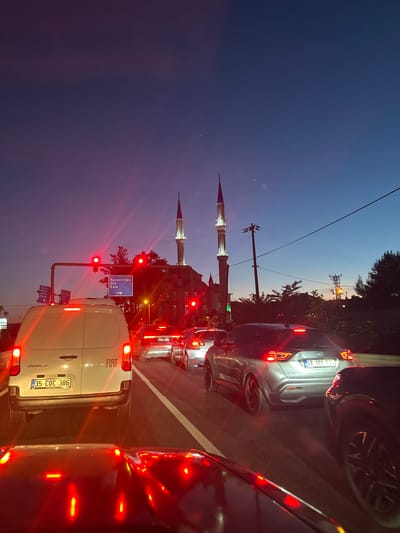 The image depicts a street scene in Ula, Turkey at dusk. A line of cars is stopped at a traffic light, with red tail lights illuminated. Dominating the left side of the frame are two minarets belonging to a mosque, a common architectural feature in Turkey. A traffic sign indicates directions to Karabörtlen and Kale. The sky transitions from dark blue to a lighter twilight hue. Power lines run across the upper portion of the sky. The cars closest to the camera obstruct a clear view of the background, giving a sense of congestion and urban life. License plates are visible on the vehicles.