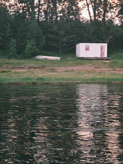 The image depicts a body of water in the foreground, with ripples reflecting the muted light of what appears to be dusk or dawn. In the middle ground, a grassy bank leads to a line of trees. On the bank, there is a small, white rectangular structure with a window and a door, resembling a shed or small cabin. A light-colored boat is also visible on the grass near the trees. The background is dominated by a dense forest, with the tops of the trees suggesting a slightly overcast sky. There is no visible evidence of human activity or any identifiable landmarks specific to the city of Yukseevo, Russia. The overall scene conveys a sense of rural isolation and tranquility.