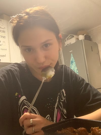 The image is a close-up, slightly blurred, vertical photograph of a young person holding a spoon with food to their mouth. The person has brown hair styled in a bun and is wearing a dark t-shirt with a graphic design featuring abstract shapes and colors. They are also wearing rings on their fingers. The background is out of focus but appears to be an indoor setting, possibly a kitchen or dining area, with some kitchen appliances visible. A black plate with food is visible on the right side of the frame. The lighting suggests it might be daytime, but the overall mood is casual and personal. Some Cyrillic text is partially visible on a white surface in the bottom left corner of the image, but it is mostly obscured and illegible. The photograph appears to be a selfie or a casual shot taken by someone else.