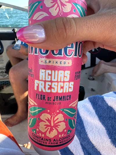 A close-up, high-angle view depicts a light-skinned hand, exhibiting manicured nails with an iridescent pink and clear gradient, gripping a condensation-covered Modelo Spiked Aguas Frescas 'Flor de Jamaica Hibiscus' can. The can is pink with light pink hibiscus flower and green leaf patterns. Visible text includes "MODELO", "SPIKED", "AGUAS FRESCAS", "FLOR DE JAMAICA", "HIBISCUS", "CONTAINS ALCOHOL", and "IMPORTED FROM MEXICO". The scene is aboard a white boat on a body of water, consistent with Folsom Lake near Granite Bay, United States. In the background, partially visible are the bare legs and feet of at least two other individuals, one person appearing to wear blue shorts. Elements of the boat's interior and controls are discernible on the left. A blue and white striped towel occupies the lower right, and a white towel with an orange stripe is in the lower left. The environment is outdoors under daylight conditions, with choppy water visible behind the boat.