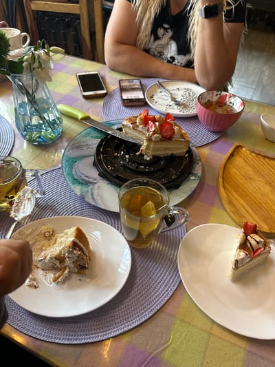 The image captures an indoor table setting, likely during an evening dessert or tea gathering, as indicated by a phone displaying "19:23." The table is adorned with a multicolored checkered tablecloth in shades of yellow, purple, and green, and two purple braided placemats are visible.
The main subjects are various food and drink items. Several slices of cake, featuring cream, strawberries, and possibly chocolate drizzle, are served on white oval plates. A larger, partially consumed cake rests on a patterned plate with a black base. Two transparent glass mugs filled with tea are present; one contains a lemon slice, and the other has a tag reading "Mickey Tea."
At least two individuals are implied. One person's arm, adorned with an Apple Watch, is visible on the left, alongside blonde, frizzy hair and part of a dark top. Another hand is seen in the bottom right, holding a spoon, suggesting active participation in eating. Two smartphones are placed on the table, one displaying the time.
Other objects include a green-handled knife, various white and pink cups or mugs, an empty pink bowl with an interior floral pattern, a white plate with an animal print design, a wooden serving board, and a light blue glass vase holding white flowers. The arrangement suggests a casual and pleasant social occasion in Matveevskoe, Russia.