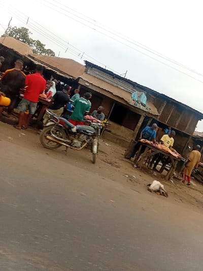 The image depicts a roadside market scene in Itobe, Nigeria, under an overcast sky. A dark, paved road occupies the foreground and left side. Adjacent to the road, on an unpaved dirt shoulder, a dark motorcycle with a chrome exhaust pipe is parked facing right.
Multiple adult males are present in the mid-ground. Several men are positioned around wooden tables that display cuts of raw meat. One man in a red shirt and blue shorts stands on the far left, holding a yellow container. Another man in a green t-shirt with white text stands near the center. To the right, a white, partially processed animal carcass, possibly a goat or sheep, lies on the dirt ground between the road and a wooden table. Further right, a man in a yellow t-shirt stands behind a table also laden with raw meat, and another man in a long, light brown garment is visible.
In the background, a series of makeshift structures with corrugated metal roofs line the street. These buildings appear to be constructed from materials such as concrete blocks, wood, and corrugated metal. Overhead wires are visible against the sky. Sparse trees are present in the upper left background. The overall scene suggests an active open-air meat market.