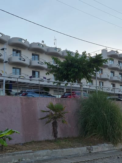 The image depicts the exterior of a hotel in Nea Kallikratia, Greece, with a street and sidewalk in the foreground. The hotel is a multi-story building with a unique architectural design featuring curved balconies with oval windows. Several cars are parked in front of the hotel. Vegetation, including a palm tree and tall grass, is visible in the foreground, suggesting a warm climate. The sky is overcast, indicating potential for changing weather. The scene is tranquil and appears to capture a moment in the late afternoon or early evening.