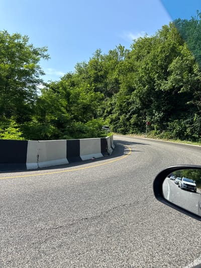 An asphalt road curves through a densely wooded area under a clear blue sky. A concrete barrier, painted with alternating black and white sections, lines the left side of the road. A yellow line marks the edge of the road. In the distance, a red and white road sign indicates a turn. Reflected in the side mirror of a vehicle, several cars are visible driving on the road ahead. The scene appears to be located in Dederkoi, Russia.