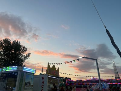 The image captures a scene at an amusement park in Golubitskaya, Russia, during sunset. The sky is a gradient of blue, with streaks of pink and orange from the setting sun illuminating fluffy clouds. In the foreground, to the left, a ride called "HAMMER TURBO 360°" is visible, adorned with colorful LED lights. To its right, a ticket booth labeled "КАССА" with a crown decoration sits under a string of colorful triangular flags. Further back, various amusement rides with flashing lights are visible, including what appears to be a mini Eiffel Tower replica in the distance. A tall lighting pole with a lamp extends upwards, and a thick rope is tautly stretched against the sky. Several people are present, some queuing at the ticket booth and others standing near the rides, engaged in their evening amusement. A large tree with dark green foliage is on the far left, silhouetted against the vibrant sky.