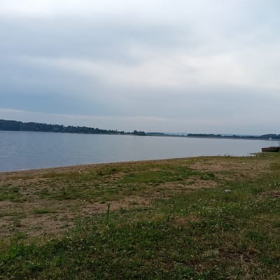 The image depicts a tranquil lakeside scene. The main subject is a wide, calm body of water, likely a reservoir, stretching into the distance. The water is a muted blue-grey, reflecting the overcast sky. A sandy shoreline occupies the foreground, with patches of dry grass and some low-lying green vegetation. In the mid-ground, a tree-lined shore creates a dark, dense silhouette against the water. A small, reddish-brown boat is beached on the grassy area to the right of the frame. In the far distance, the treeline continues, with hints of buildings and structures visible along the horizon. The sky is heavily clouded, suggesting an overcast or possibly pre-stormy day. The overall impression is one of quietude and natural beauty, with a sense of solitude. The time of day appears to be late afternoon or early evening, given the soft, diffused light. The location cues, such as the type of vegetation and the presence of a lake, are consistent with a rural or semi-rural setting. No people are visible in the image, and there is no visible text.