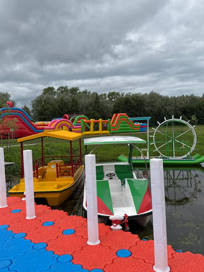 This image depicts an outdoor recreational area on a cloudy day, likely in Bruzdeilynas, Lithuania, featuring a body of water and various attractions. In the foreground, a vibrant red and blue modular floating dock with white vertical bollards leads to two small boats docked side-by-side. The boat on the left is yellow with a red-framed canopy, while the boat on the right is white with green and red accents and a steering wheel.

In the background, several inflatable play structures, including a large red and blue slide and other colorful obstacles, are visible on a grassy area. Adjacent to the inflatables and on the water, there is a large green Ferris wheel-like structure mounted on a raft. Dense green trees form a backdrop to the scene. The water is dark and appears to have some aquatic plants floating on the surface. The sky is overcast with gray clouds, suggesting the possibility of rain. No people are clearly visible in the immediate vicinity of the boats or dock.