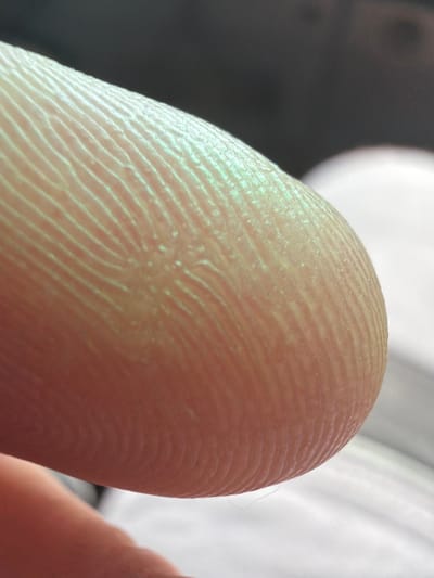 The image is an extreme close-up of a human fingertip. The subject is a person's finger, with visible fingerprint ridges. The skin appears pale with a slightly yellowish or greenish tint due to the lighting. The background is out of focus, with a dark, shadowed area on the left and a blurred bright area on the right, suggesting artificial lighting or a bright light source out of frame. There is a hint of another finger or hand in the lower right corner, also out of focus. The activity appears to be a static shot, focusing on the texture of the skin and the detail of the fingerprint. There are no visible location cues, time of day indicators, weather conditions, or emotions conveyed. No text is visible in the image.