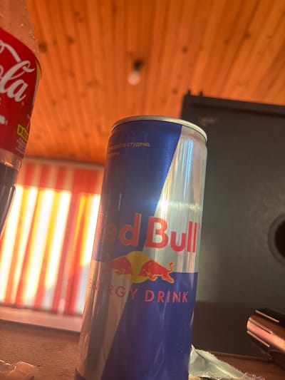 The image displays a close-up, angled view of a Red Bull energy drink can. The can is predominantly blue with silver accents and features the iconic Red Bull logo with a red bull and sun. Text on the can is partially visible, including "Red Bull" and "ENERGY DRINK," as well as some text in what appears to be Cyrillic script. To the left and bottom of the Red Bull can, a portion of a Coca-Cola bottle is visible, also red and white. The background consists of a warm-toned wooden surface, likely paneling, which suggests an indoor setting. In the upper right portion of the image, a metallic object with parallel grooves is visible, possibly part of a lamp or another appliance. There are vertical stripes of light and shadow in the background, creating a textured visual. The lighting in the image is somewhat diffuse, with reflections visible on the can, indicating it's likely daytime or well-lit indoors. The overall impression is a casual, perhaps domestic or recreational environment, given the presence of beverages. The location context is Zlataritsa, Bulgaria.