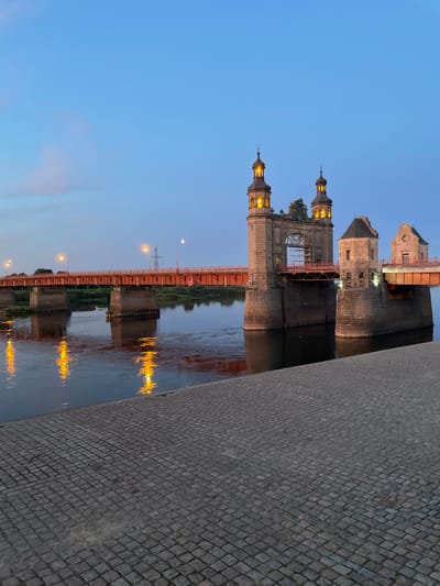 A long shot shows a wide, tranquil river with reflections of yellow lights from a red-railed bridge. The bridge has stone pillars and two towers with illuminated roofs. The sky is a clear, pale blue with a hint of pink and a few wispy clouds. In the foreground, a cobblestone walkway leads to the riverbank. The scene is set in Sovetsk, Russia, with the bridge being the Bridge of Freedom. There are no people or animals visible in the image. The overall atmosphere is peaceful and serene, suggesting dusk or early dawn.