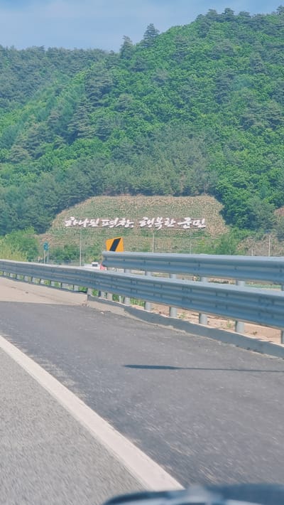 The image shows a view from a vehicle traveling on a road in Yongpyeong-myeon, South Korea. A guardrail runs along the side of the road, separating it from a wooded area. The guardrail is metal and appears to be in good condition. Beyond the guardrail is a hillside covered in trees. There is a sign, partially obscured by the guardrail, that has yellow and black stripes. Adjacent to the road and sign, there is a row of white characters on the hill. The road surface is asphalt, with a white line marking the edge of the lane. The sky is visible in the upper left corner of the frame.