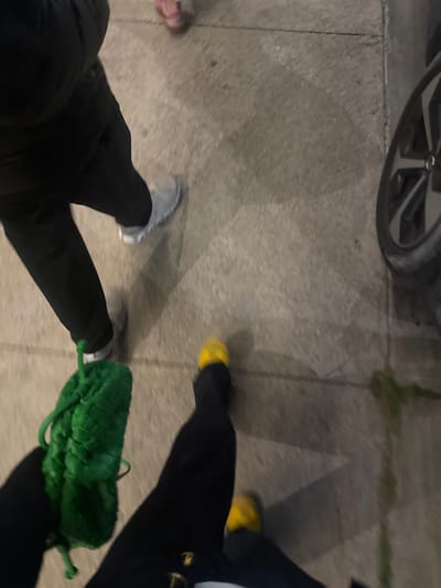 The image depicts the lower extremities of at least three individuals walking on a light grey concrete sidewalk in La Purísima, Mexico. In the left foreground, a person wears dark trousers and a grey athletic shoe on their visible right foot, holding a distinct green woven drawstring bag in their left hand. Immediately to their right, another individual is seen wearing dark trousers and bright yellow shoes. In the upper background, a small foot in a pink shoe is partially visible, suggesting a child is also present. All subjects appear to be in motion. The concrete pavement exhibits visible seams and minor debris. Elongated shadows are cast across the ground, indicating a light source. On the far right, a section of a dark vehicle tire with a silver multi-spoke wheel rim is visible, with sparse green vegetation at the base of the tire.