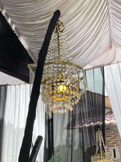 The image depicts an indoor or semi-outdoor event space located in Matangbaroh, Indonesia. A prominent golden-framed chandelier hangs centrally, adorned with multiple clear and yellowish crystal-like pendants. An incandescent bulb within the chandelier emits a warm, yellow light. The chandelier is suspended from a thick, vertical support pole wrapped in black fabric, extending from the upper-left towards the bottom-center.

The ceiling consists of draped fabric, with white pleated material dominating the upper-center and right, and a section of black fabric forming the upper-left. A ruffled border separates the white and black ceiling fabrics. In the background, translucent black drapes are visible, layered with white drapes on the far right. A second, smaller chandelier-like decoration, primarily white, is partially visible to the left and slightly behind the main chandelier. Towards the lower right, a brick wall with a small window-like opening and a section of a reddish-brown tiled roof are visible. The bottom of the image shows a dark, cylindrical object and faint green foliage. No individuals are present in the scene.