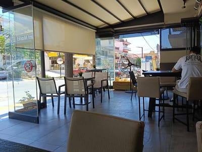 The image depicts an outdoor seating area of a cafe or restaurant in Nea Kallikrateia, Greece. The scene is viewed from the perspective of someone sitting at a table, looking towards the street. The foreground is dominated by tables and chairs, with one person, a male, visible from the back, wearing a white t-shirt with graphics. He is seated at a table, facing away from the camera. The seating area is covered by a retractable awning with a light-colored fabric. Glass partitions separate the seating area from the sidewalk and street.

Through the glass, we see a street with parked cars, a red car, and buildings. Signs are visible, including one with Greek text and another advertising "BEAN COFFEE SHOP." A large industrial-style fan is positioned outdoors, in front of the glass. Plants in pots are also visible, both inside and outside the seating area. The overall impression is a sunny, urban outdoor cafe environment.