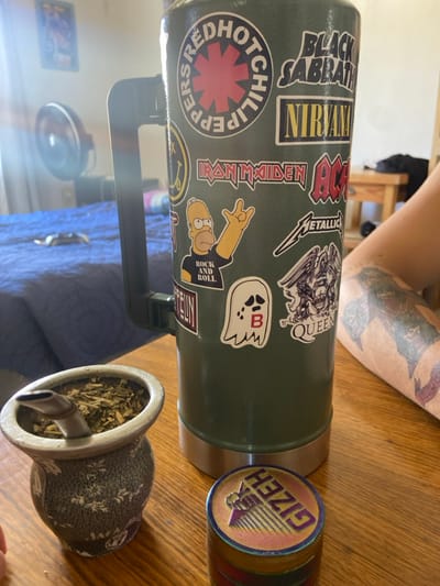 A green thermos decorated with rock band stickers like Black Sabbath, Nirvana, AC/DC, Metallica, and Queen sits on a wooden table. A colorful, multi-tiered grinder labeled "GIZEH" rests nearby. Next to the thermos is a grey, ceramic mate gourd filled with yerba mate and a silver bombilla. Part of a blue blanket and an arm with a large tattoo are visible in the background.