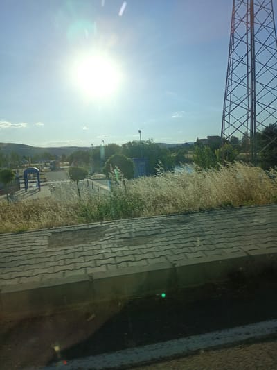 This media file captures an outdoor scene with a bright sun in a clear blue sky, suggesting it is daytime and likely sunny weather. In the foreground, a paved surface with a tiled pattern is visible, with some dry, wispy vegetation growing in patches and along the edges. A portion of a tall, lattice-like metal tower, possibly a communication tower, dominates the right side of the frame, stretching upwards into the sky.
The middle ground reveals a park-like area with a blue archway structure, some trees, and paved pathways. There are also streetlights visible, indicating a public space. Beyond the park, a gently rolling landscape with hills and scattered trees can be seen, with some buildings subtly integrated into the terrain. The overall impression is of a somewhat rural or semi-urban environment.
There is no visible text in the image. The scene appears to be tranquil and possibly depicts a leisure or public space. The time of day is indicated by the strong sunlight and the long shadows cast by some of the objects, suggesting it is either morning or late afternoon. The location context provided places this scene in Bahşılı Belediyesi, Turkey.