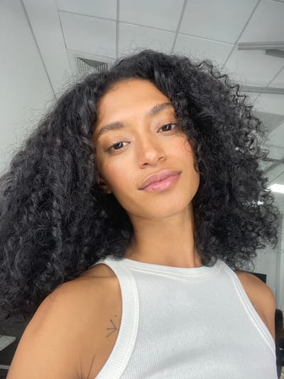 A medium-angle, close-up shot depicts a woman wearing a white tank top. She has dark, curly hair and light skin. The background is a white tiled wall and ceiling. A small tattoo is visible on her arm. The location appears to be indoors, but no landmarks or identifying information suggest that it is in Porto Alegre, Brazil.