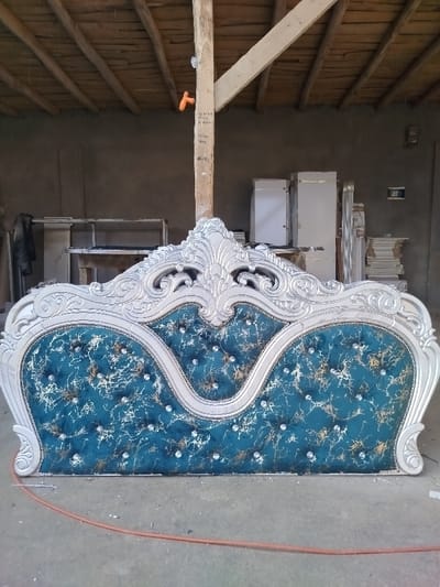 A luxurious, ornate headboard stands prominently. Its frame is painted in shimmering silver, featuring elaborate floral and scrollwork details. The upholstered section is deep teal, with a marbled pattern in gold and silver. It's tufted with sparkling crystal buttons. The headboard sits on a concrete floor in what appears to be a workshop or storage area, with wooden beams visible above, and various construction and storage materials surrounding it.