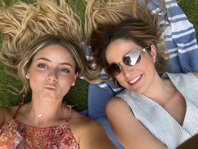 Two women are lying on the ground, facing the camera. The woman on the left wears sunglasses and a striped shirt. She is resting on a striped towel. The woman on the right wears a patterned top and a silver necklace. Their hair fans out on the green grass behind them. The photo appears to have been taken from directly above them.