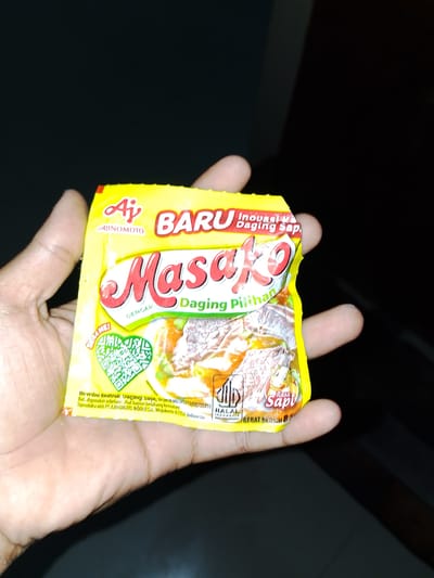 A person's hand holds a yellow packet of "Masako" brand seasoning. The packet has red lettering for "Masako" and other text in white and green. The product is described as "Daging Pilihan" (Selected Meat) and "Rasa Sapi" (Beef Flavor). A QR code is visible on the packet. The background is dark and out of focus. The location of the image is stated as Sendangrejo, Indonesia, on the packaging.