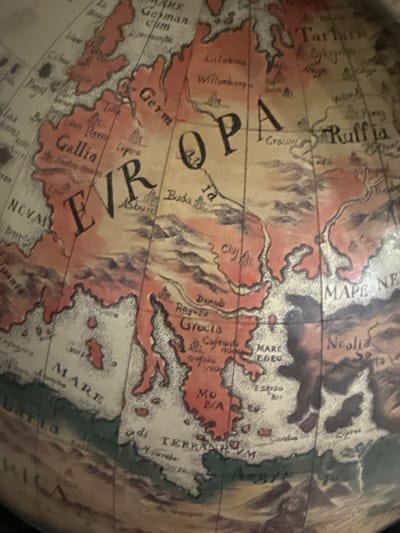 This is a close-up, angled shot of a section of an antique globe, focusing on Europe and parts of Africa and Asia. The globe's surface is detailed with painted landmasses in shades of pink and brown, and bodies of water in a light beige color with dotted textures. The lines of latitude and longitude are visible, dividing the sphere.
The map displays various place names in a stylized, serif font. Visible text includes "Ruffia," "EUROPA," "Germ," "Gallia," "MARE EGEO," "MARE," and "TERRA." Other smaller place names and labels are scattered across the map, some of which are partially legible. The overall impression is of historical cartography, with a hand-drawn aesthetic. The lighting appears to be natural, casting subtle shadows and highlighting the texture of the globe's surface. There are no people or animals depicted. The setting is likely an indoor space where the globe is displayed or stored.