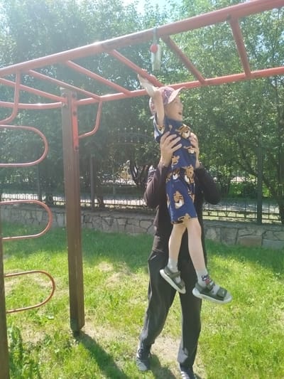 In an outdoor setting, a person supports a young child who is hanging from a red monkey bar structure. The child, wearing a patterned t-shirt, shorts with a tiger print, a cap, socks, and sandals, is being held up by an adult who is wearing dark pants and a dark shirt. The monkey bars are part of a playground apparatus set against a backdrop of green trees and a blue sky. The ground is covered in grass, with a stone wall and a decorative fence visible in the background. The scene appears to be a public park or playground.