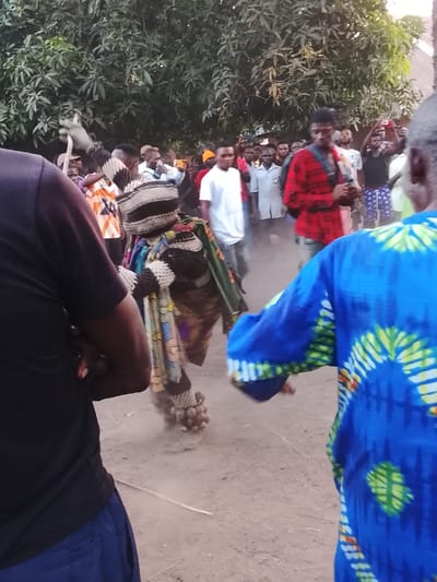 The image depicts an outdoor cultural event in Tabu, Nigeria, centered around a masked figure. The scene is on a dirt ground surface, with lush green foliage from trees visible in the background.
In the foreground, two individuals are partially visible from behind. On the left, a person wears a black short-sleeved top and dark blue trousers. On the right, another person wears a vibrant blue long-sleeved garment with yellow and green tie-dye patterns. Both appear to be observing the central activity.
The central subject is an elaborately costumed masked figure, facing leftward, kicking up dust from the ground. This figure is adorned with a tall, striped headpiece (black and white), multiple layers of patterned fabric in varying colors (brown, green, multi-colored geometric designs), and a cluster of light-colored, spherical objects attached to its lower legs, likely rattles or traditional regalia.
Behind the central figure, a crowd of approximately 20-30 spectators, predominantly male, observes the event. Several individuals within the crowd are holding up mobile phones, apparently recording or photographing the spectacle. Notable figures in the crowd include a man in a white t-shirt and another in a red and black plaid shirt. Dust is visibly suspended in the air around the central figure due to movement. A thin, light-colored stick lies on the ground in the lower middle of the frame.