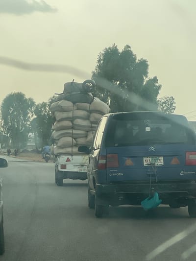 The image depicts a road scene in Kasuwan Magani, Nigeria. A white truck overloaded with sacks is followed closely by a blue Citroen Evasion. The Citroen's license plate reads "MKA-697ZE" and the truck's plate reads "DKA-787ZG." Trees line the road. A person wearing blue clothing stands near the truck. The sky is overcast.