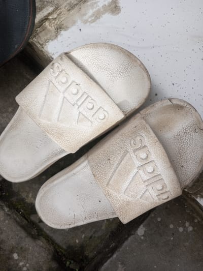 Two worn, white Adidas slide sandals sit on a textured surface. The sandals show signs of wear, with dirt and cracks visible on the straps and soles. The Adidas logo is prominently displayed on each strap. The background consists of a mottled surface with concrete and brick-like patterns.