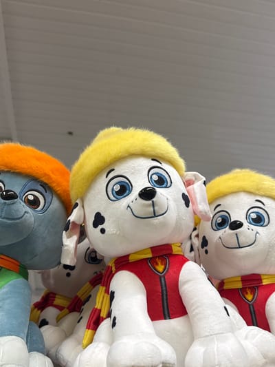 This media file is a close-up, vertically oriented photograph of several stuffed animal dogs stacked on top of each other. The dogs appear to be characters from the Paw Patrol franchise, with distinctive white fur, large blue eyes, and black spots. Each dog is wearing a red vest with yellow trim and a yellow scarf. The top two dogs are clearly visible, with their faces and torsos dominating the frame. The dog in the middle has a yellow fluffy collar or hood around its head. Below these two, a portion of a third stuffed dog is visible, this one is blue-grey and has an orange fluffy collar or hood. The background is a plain, ribbed, light gray surface, possibly a wall or a corrugated metal surface. There is no discernible text visible in the image. The lighting is bright and even, suggesting it was taken indoors in a well-lit area, possibly a store or a playroom. The overall impression is one of a collection of children's toys. The location context of Ivanivka, Ukraine, is not visually apparent from the image itself.