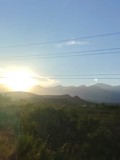 This image depicts a serene, natural landscape at sunrise or sunset, likely in a mountainous region of Kyrgyzstan. The sun, a brilliant orb of light, is low on the horizon and partially obscured by hills, casting a warm glow across the scene. The sky transitions from a bright, hazy yellow near the sun to a clear, pale blue at the top of the frame. Three thin, dark horizontal lines, presumably power lines, stretch across the sky, subtly dividing the frame.

The foreground is dominated by lush green vegetation, including trees and tall grasses, which are rendered in shadow due to the low angle of the sun. Beyond this verdant area, rolling hills rise, their shapes softened by the hazy light. In the middle ground, silhouetted against the bright sky, are distinct mountain ranges with jagged peaks. The overall impression is one of tranquility and natural beauty, enhanced by the soft, diffused light and the vastness of the landscape. The time of day is clearly indicated by the sun's position and the quality of light. There are no visible people, text, or specific activity taking place, suggesting a moment captured in stillness. The environment appears remote and undeveloped, consistent with a rural or mountainous setting.